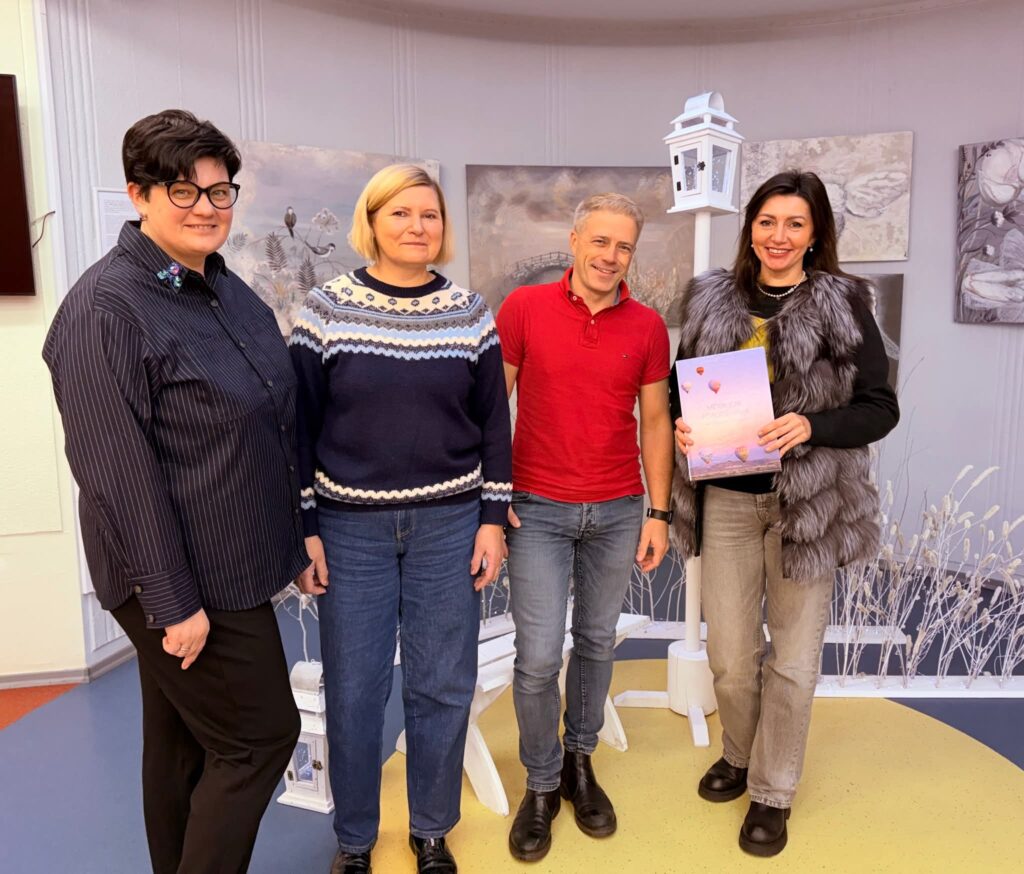 Keturi suaugusieji pozuoja bibliotekos patalpoje prie parodos ekspozicijos: trys moterys ir vyras stovi greta, viena moteris rankose laiko knygą. Fone matyti šviesių tonų meno darbai ir dekoratyvinis žibintas.