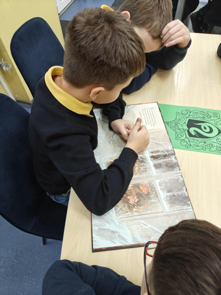 Trys vaikai sėdi prie stalo ir kartu skaito bei apžiūrinėja didelę iliustruotą knygą. Vienas berniukas pasilenkęs virš atversto puslapio, rodo pirštu į tekstą ar paveikslėlius, kiti du stebi. Ant stalo matyti žalia kortelė su stilizuotu akies simboliu. Bibliotekos aplinka – šviesus stalas, kėdės, tvarkinga mokymosi erdvė.