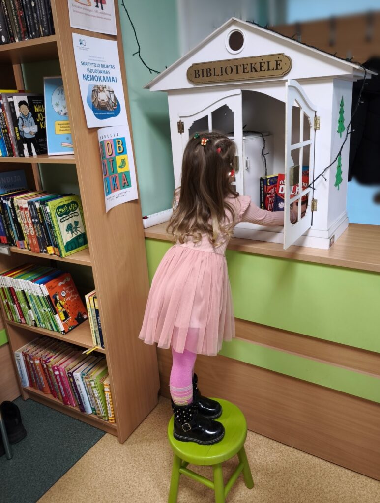 Maža mergaitė, stovinti ant žalios taburetės bibliotekoje, renkasi knygą iš mažo, balto namelio formos knygų spintelės su užrašu „Bibliotekėlė“. Ji vilki rožinę suknelę, rožines pėdkelnes ir juodus batus. Aplink matyti knygų lentynos su vaikiškomis knygomis, ant sienos – informaciniai plakatai.