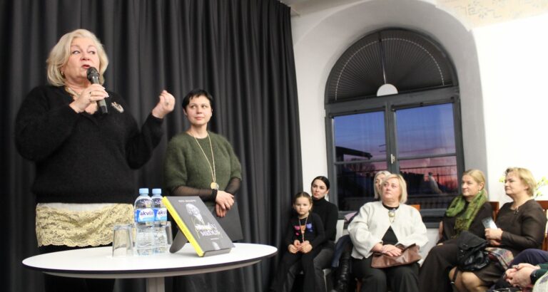 Renginio metu Edita Mildažytė kalba į mikrofoną, šalia jos stovi kita dalyvė, o priešais sėdi klausytojai. Ant stalo – pristatoma knyga ir vandens buteliukai.