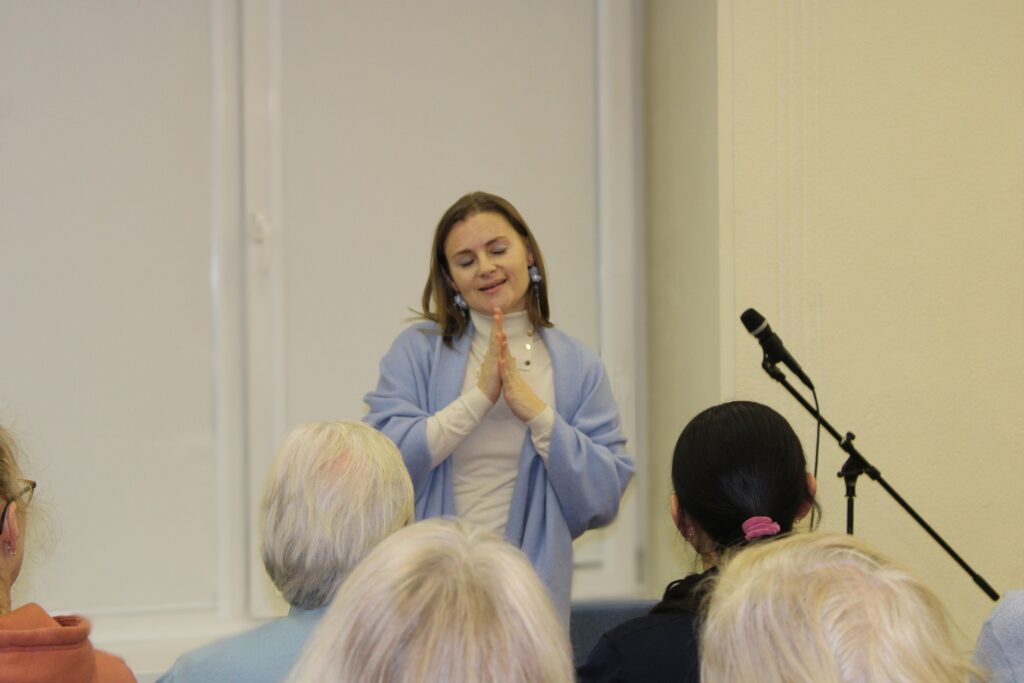 Moteris stovi prieš auditoriją ir kalba, sudėjusi rankas maldos poza. Ji vilki šviesius drabužius ir mėlyną skarą. Priekyje matyti sėdintys klausytojai, šone – mikrofonas ant stovo. Aplinka rami, primenanti paskaitą ar susitikimą.