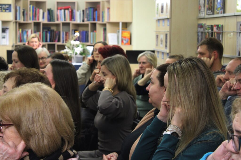 Žmonių grupė sėdi auditorijoje ar bibliotekoje ir atidžiai klausosi užsiėmimo, dauguma dalyvių rankomis prisilietę prie ausų ar veido.