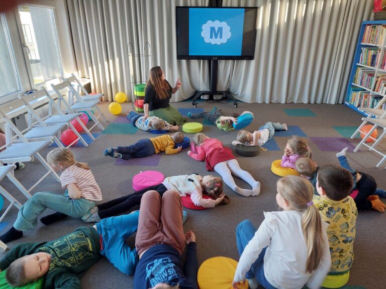 Gulintys ant grindų ir sėdintys vaikai su suaugusiąja vadove bibliotekos erdvėje