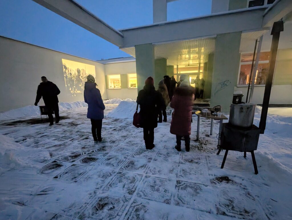 Žiemos vakaras pastato kieme: sniegu padengtoje erdvėje vaikšto žmonės su žieminiais drabužiais, ant sienos rodomas archyvinis vaizdo įrašas, o šalia stovi krosnelė su karštais gėrimais.