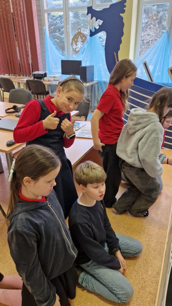 Grupė pradinių klasių mokinių klasėje: keli vaikai klūpo prie lentos ar stendo, o viena mergaitė su raudonais marškinėliais ir tamsia uniforma atsisukusi į kamerą rodo pakeltus nykščius. Fone matyti kompiuteriai, langai su žiemiškomis dekoracijomis, mėlynos užuolaidos ir vikingų laivo maketo priekis (laivo nosis).