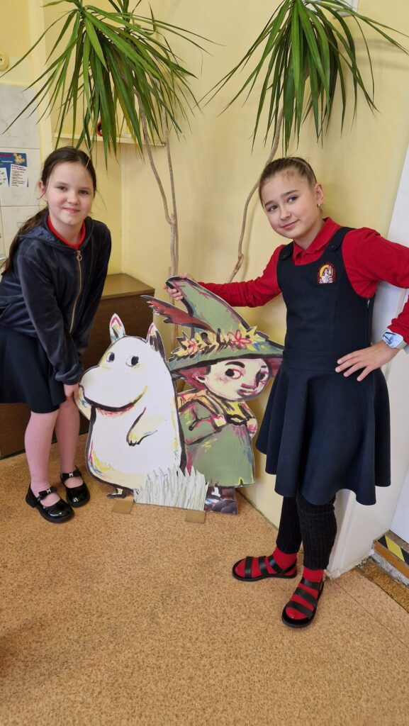 Dvi mergaitės stovi klasėje šalia kartoninių Muminuko ir Snusmumriko figūrų iš Tovės Jansson knygų – viena laiko leteną balto, į begemotą panašaus Muminuko, kita atsirėmusi į berniuko su skrybėle ir plunksna figūrą; už jų matyti aukšti kambariniai augalai.