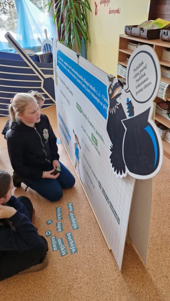 Du vaikai klūpo ant grindų ir dėlioja žodžių korteles prie mokomosios lentos su varnos iliustracija ir užduotimi sudėti trūkstamas eilutes.