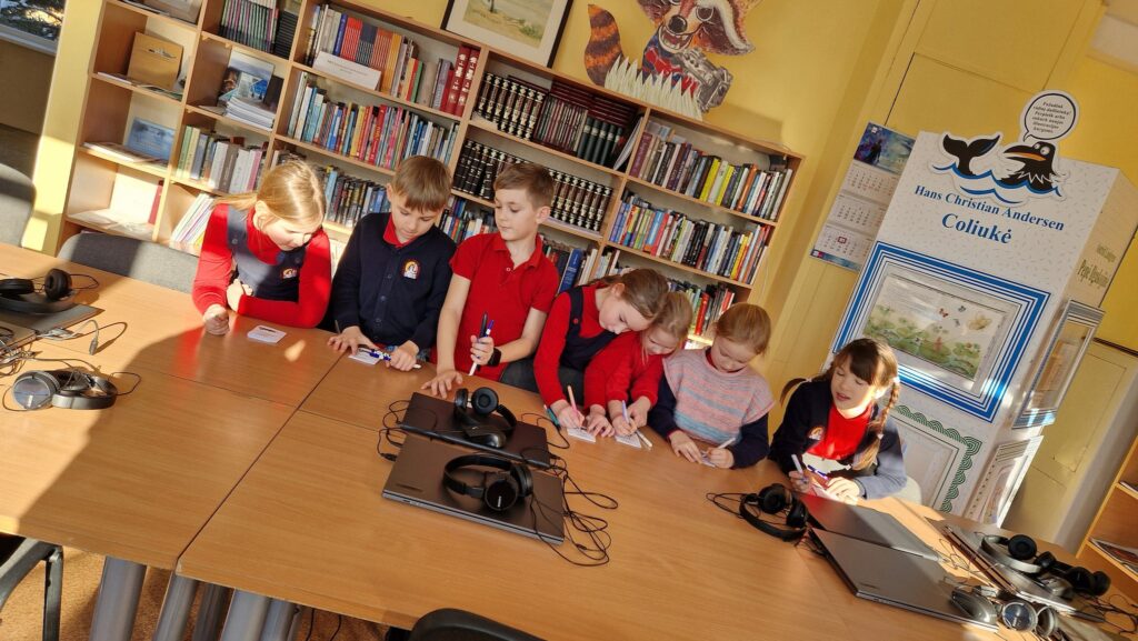 Šeši vaikai bibliotekoje sėdi prie stalo ir rašo ant kortelių, o ant stalo padėti nešiojamieji kompiuteriai bei ausinės; fone matyti knygų lentynos ir stendas su užrašu „Hans Christian Andersen Coliukė“.
