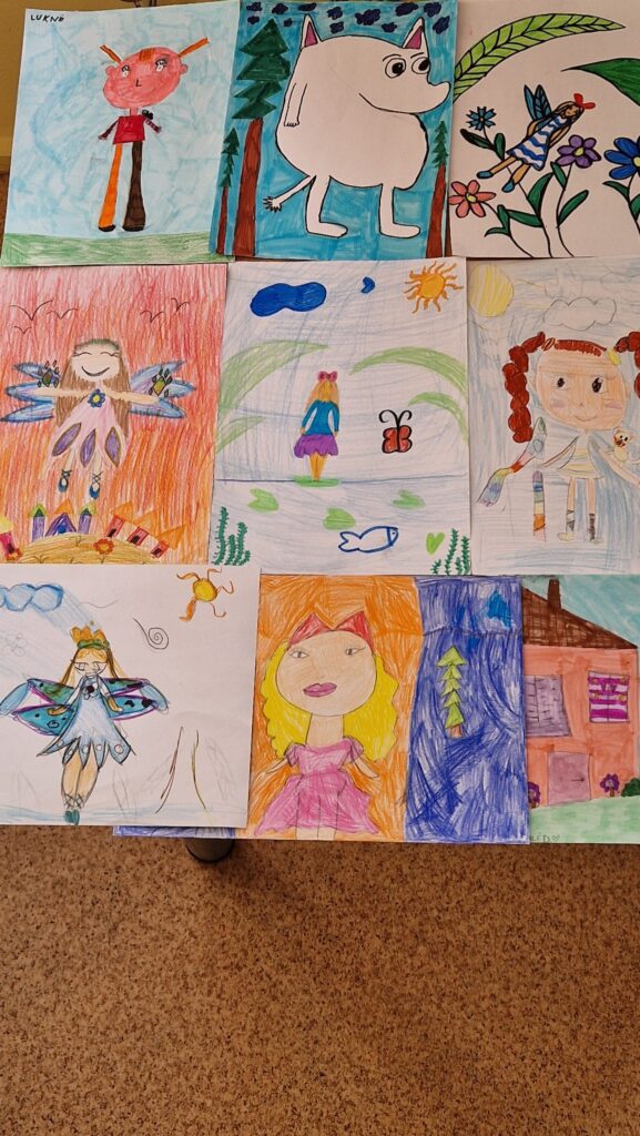 Ant stalo išdėlioti spalvingi vaikų piešiniai, kuriuose pavaizduoti pasakų veikėjai, fėjos, mergaitės, gyvūnai, namas ir gamtos motyvai – saulė, medžiai, gėlės bei dangus.
