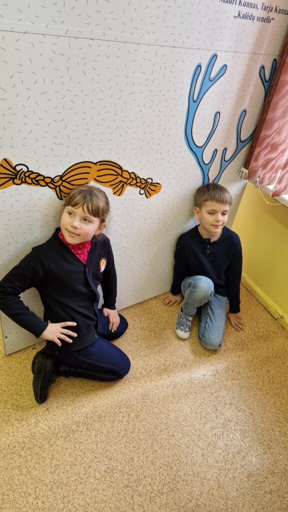 Mergaitė ir berniukas klūpo prie sienelės su pieštomis kasomis ir elnio ragais; jie pozuoja taip, kad iliustracijos sudaro žaismingą įspūdį, tarsi mergaitė turėtų ilgas kasas, o berniukas – ragus.