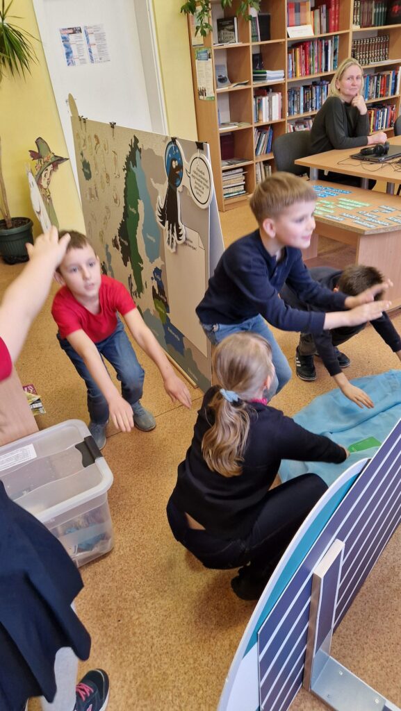 Keturi vaikai bibliotekoje pritūpę atlieka judesio užduotį prie edukacinės sienelės su žemėlapiu, o už jų prie stalo sėdi ir stebi mokytoja; aplink matyti knygų lentynos ir mokymo priemonės.