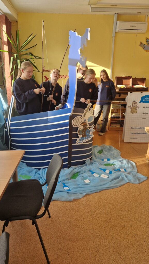 Klasėje keli mokiniai stovi prie dekoratyvaus mėlyno „Vikingų laivo“ maketo ir laiko meškeres, tarsi žvejotų. Ant grindų patiesta mėlyna medžiaga, vaizduojanti jūrą, joje išdėliotos popierinės „žinutės buteliuose“. Aplink matyti bibliotekos aplinka – kėdės, augalas, knygų lentyna ir stendas su tekstu.