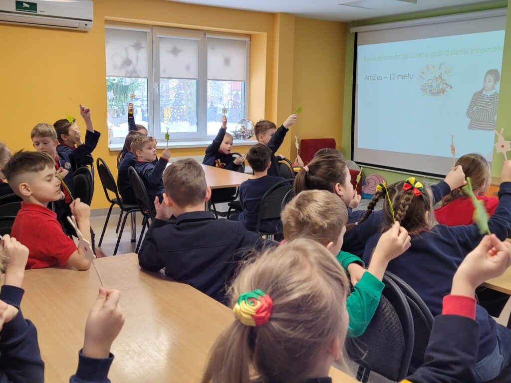 Klasėje prie stalų sėdi pradinių klasių mokiniai ir pakėlę rankose laiko mažas korteles ar ženklus. Jie žiūri į priekyje rodomą skaidrę su klausimu apie tai, kokia informacija gali būti dalijamasi internete, nurodytas amžius – 12 metų. Ant sienos matyti langai ir projektoriaus ekranas, klasėje vyrauja geltonos spalvos sienos.