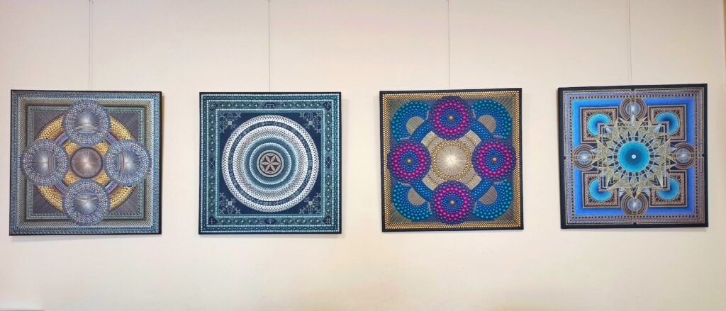 Paveikslėlyje matome keturis spalvingus geometrinio tipo paveikslus, kurie kabantys ant sienos. Kiekvienas paveikslas turi sudėtingus ir simetriškus dizainus, kuriuose naudojamos įvairios spalvos, tokios kaip mėlyna, žalia, violetinė ir auksinė. Paveikslai sukurti naudojant taškus ir linijas, sudarančius mandalų tipo motyvus, kuriuose aiškiai matosi subtilios detalės ir gilūs spalvų perėjimai. Paveikslai atrodo harmoningai išdėstyti vienas šalia kito.
