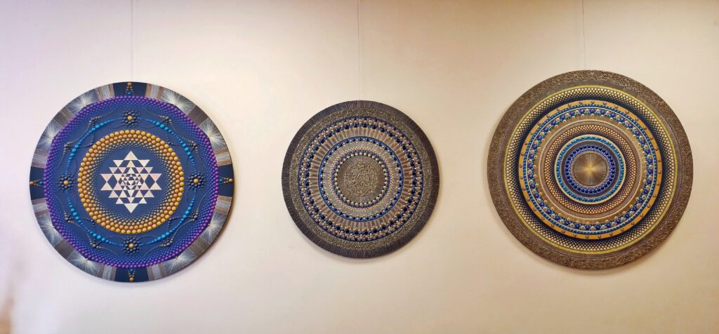 Paveikslėlyje matome tris didelius, apvalius ir spalvingus geometrinio dizaino paveikslus, kabantys ant sienos. Kiekvienas iš jų turi sudėtingą struktūrą su spalvotais taškais ir linijomis, sukuriančiomis mandalų tipo motyvus. Pirmasis paveikslas turi ryškias mėlynas, violetines ir auksines spalvas, antrasis – daugiau žemės tonų ir šviesesnį dizainą, o trečiasis – švelnesnes mėlynas ir auksines spalvas su detalėmis, panašiomis į raštus. Kiekvienas paveikslas yra unikalus, tačiau visi jie puikiai dera tarpusavyje.