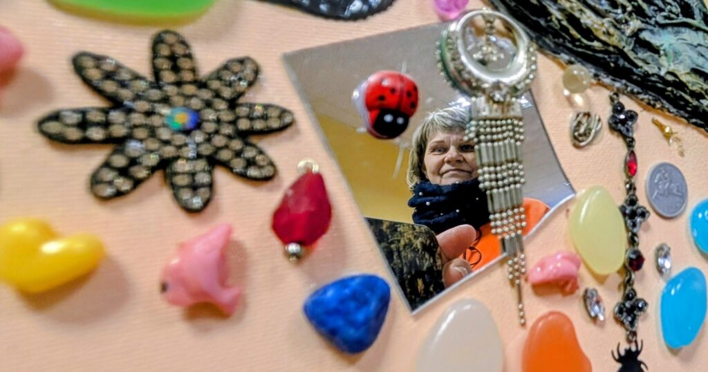 Moteris atsispindi mažame veidrodėlyje, padėtame tarp spalvingų papuošalų ir įvairių dekoratyvinių detalių ant stalo.