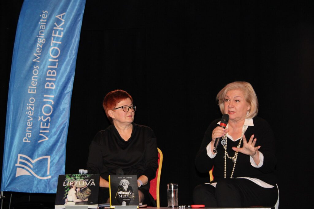 Scenoje sėdi dvi moterys. Dešinėje moteris laiko mikrofoną ir kalba, kairėje – kita moteris jos klausosi. Šalia stovi mėlyna vėliava su užrašu „Panevėžio Elenos Mezginaitės viešoji biblioteka“. Ant stalo priešais matomos knygos „Pamirštas sugrįžimas į Lietuvą“ ir „Pelynų medus“, taip pat stiklinė vandens. Tamsus fonas pabrėžia renginio aplinką.