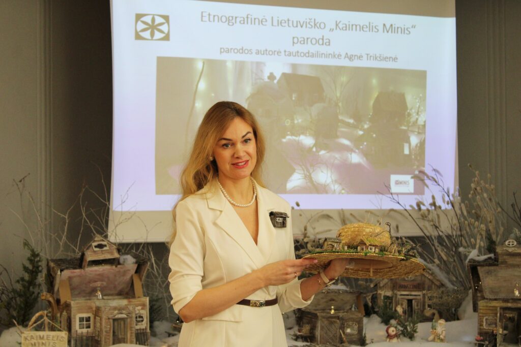 Moteris su šviesia suknele ir perlais stovi priešais pristatymo ekraną su užrašu apie etnografinę lietuviško „Kaimelis Minis“ parodą, rankose laiko šiaudinę skrybėlę, papuoštą miniatiūriniu kaimelio maketu; aplink matomi žiemiški namelių modeliai.