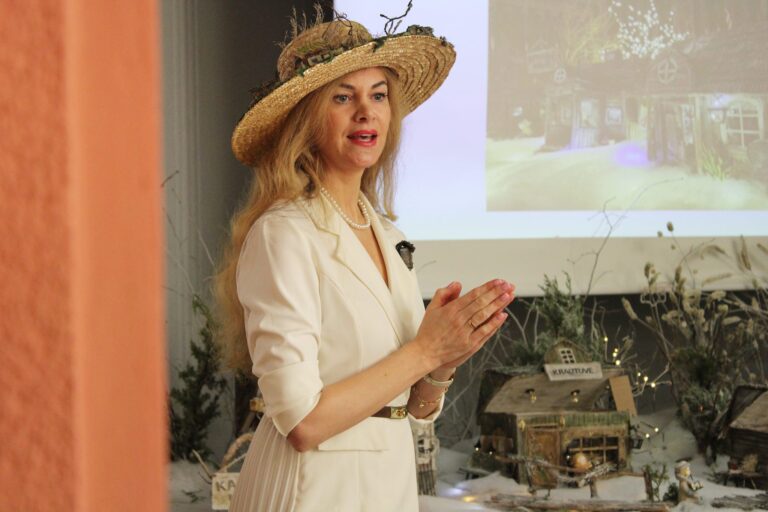 Moteris su šviesia suknele ir šiaudine skrybėle stovi prie stalo su žiemos kaimelio maketu ir kalba, rankas suglaudusi priešais save; fone matomas projektoriaus ekranas su namelio vaizdu.