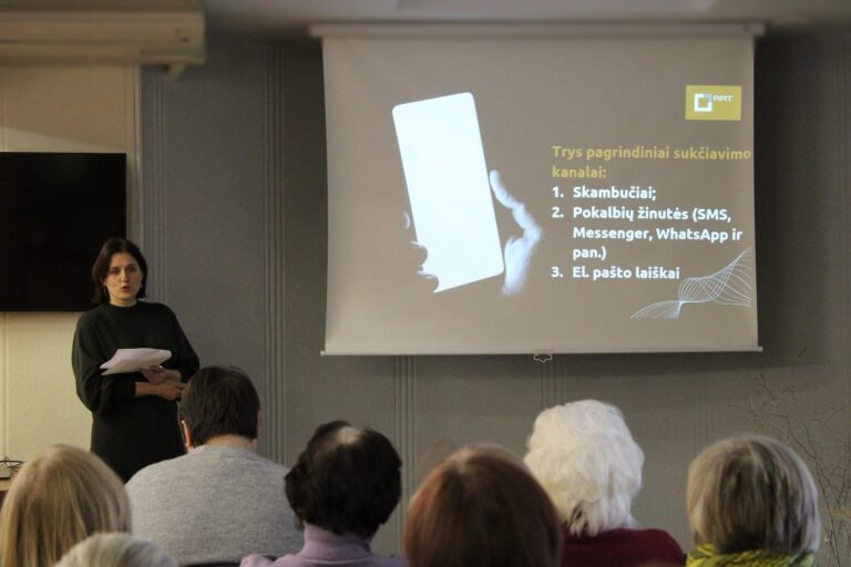 Moteris stovi prieš auditoriją ir skaito pranešimą, laikydama lapus rankoje. Už jos ekrane rodoma skaidrė su užrašu „Trys pagrindiniai sukčiavimo kanalai: 1. Skambučiai; 2. Pokalbių žinutės (SMS, Messenger, WhatsApp ir pan.); 3. El. pašto laiškai“ bei pavaizduotas rankoje laikomas mobilusis telefonas. Auditorijoje sėdi klausytojai, matomi iš nugaros.