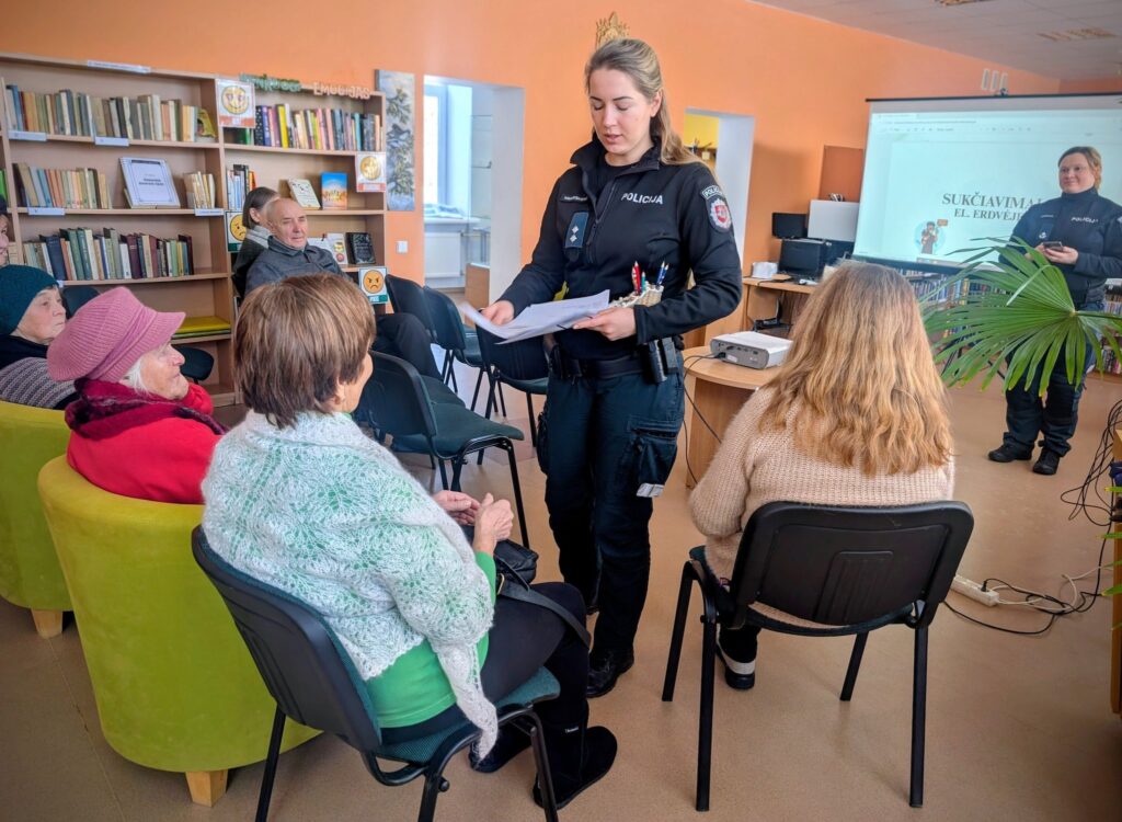 Policijos pareigūnė stovi bendruomenės susitikime ir dalina dokumentus vyresnio amžiaus žmonėms, sėdintiems kėdėse, o kitas pareigūnas rodo pristatymą ekrane patalpoje su knygų lentynomis.