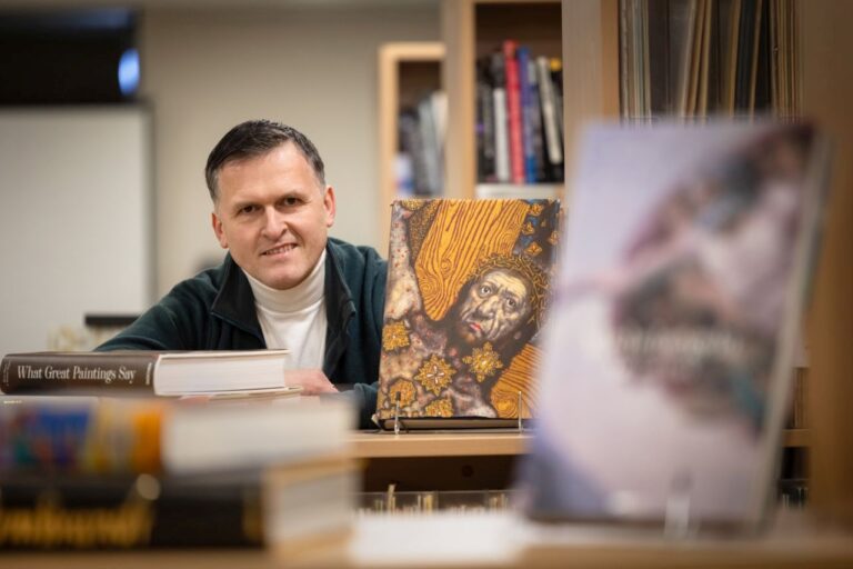 Vyras šypsosis sėdėdamas aplinkui stovinčių knygų fone