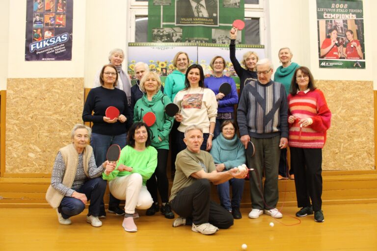 Grupinė nuotrauka patalpoje: apie penkiolika vyresnio amžiaus moterų ir vyrų stovi ir pritūpę pozuoja su stalo teniso raketėmis. Viena moteris pakėlusi raudoną raketę aukštyn, priekyje klūpantis vyras rankoje laiko kelis stalo teniso kamuoliukus. Fone – sienos su sporto plakatais ir nuotraukomis, medinė apdaila, geltonos grindys. Visi atrodo nusiteikę draugiškai, lyg sporto klubo ar treniruotės dalyviai.